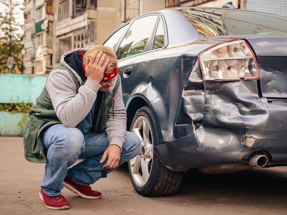 A man sits beside the damaged car post-accident, holding his head in distress.