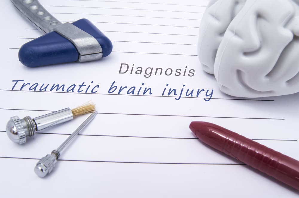 A medical diagnosis of traumatic brain injury: On a paper form lie a figure of a human brain, a blue neurological reflex hammer, a neurological needle and brush for sensitivity tests, and a ballpoint pen.