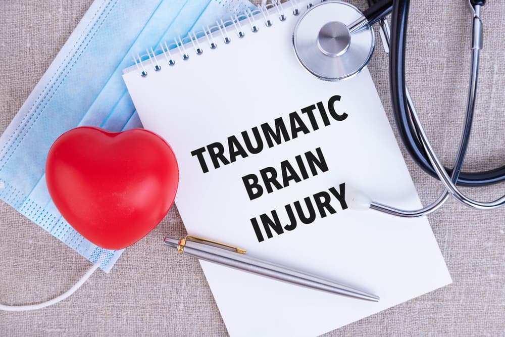 The text "Traumatic Brain Injury" is written in a notebook, accompanied by a pen, a disposable medical mask, and a stethoscope, set on a linen background.