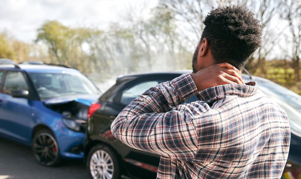 A young man stands beside a damaged car, rubbing his neck in pain from a whiplash injury after a traffic accident.