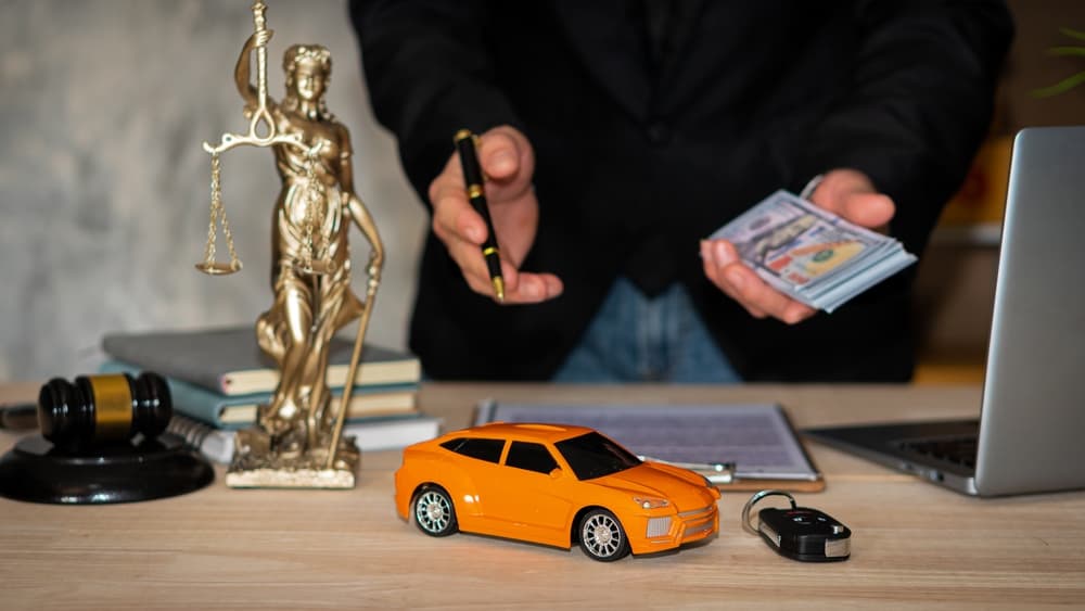 A lawyer with laptop holding contract, money, and car asset auction document, featuring a model car and the Hammer of Justice symbolizing injustice.