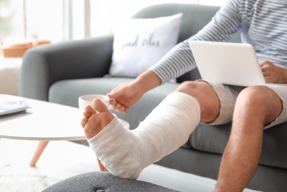 Young man with broken leg using laptop while drinking tea at home