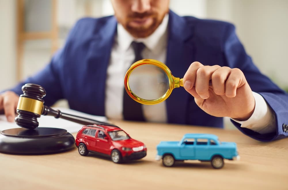A lawyer in a suit examines toy cars with a magnifying glass, symbolizing accident investigation.